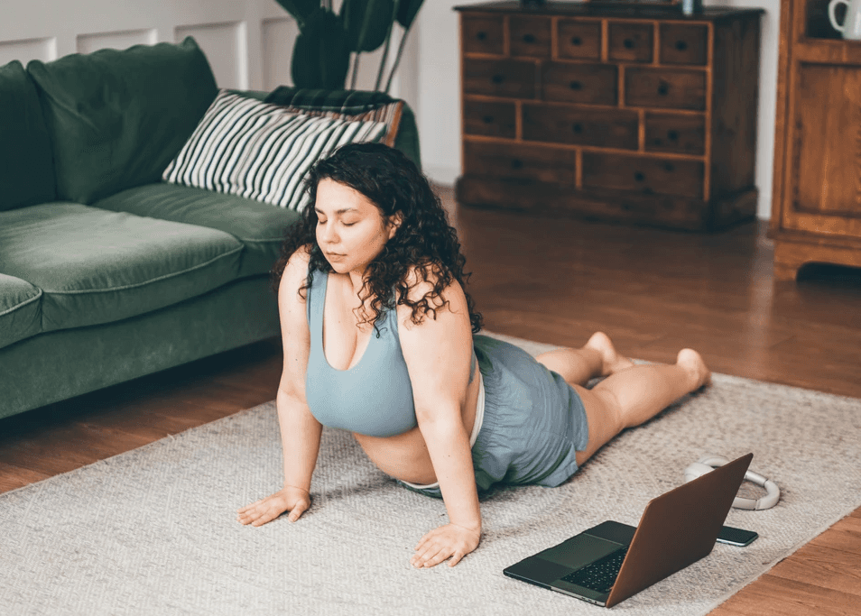 Woman stretching in upward-facing dog yoga pose in her living room with a laptop in front of her